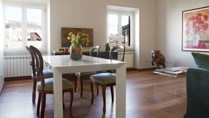 A dining area with a white table, four chairs, a vase with flowers, and a view of buildings through large windows.