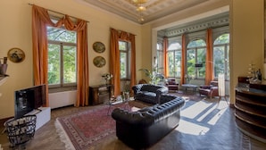 A spacious living room with a large black leather sofa, a wooden coffee table, and a red patterned rug. There are multiple windows with curtains, a fireplace, and a chandelier.