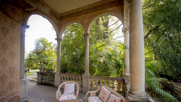 A covered outdoor seating area with two wicker chairs and a small table, surrounded by ornate columns and a view of lush greenery.