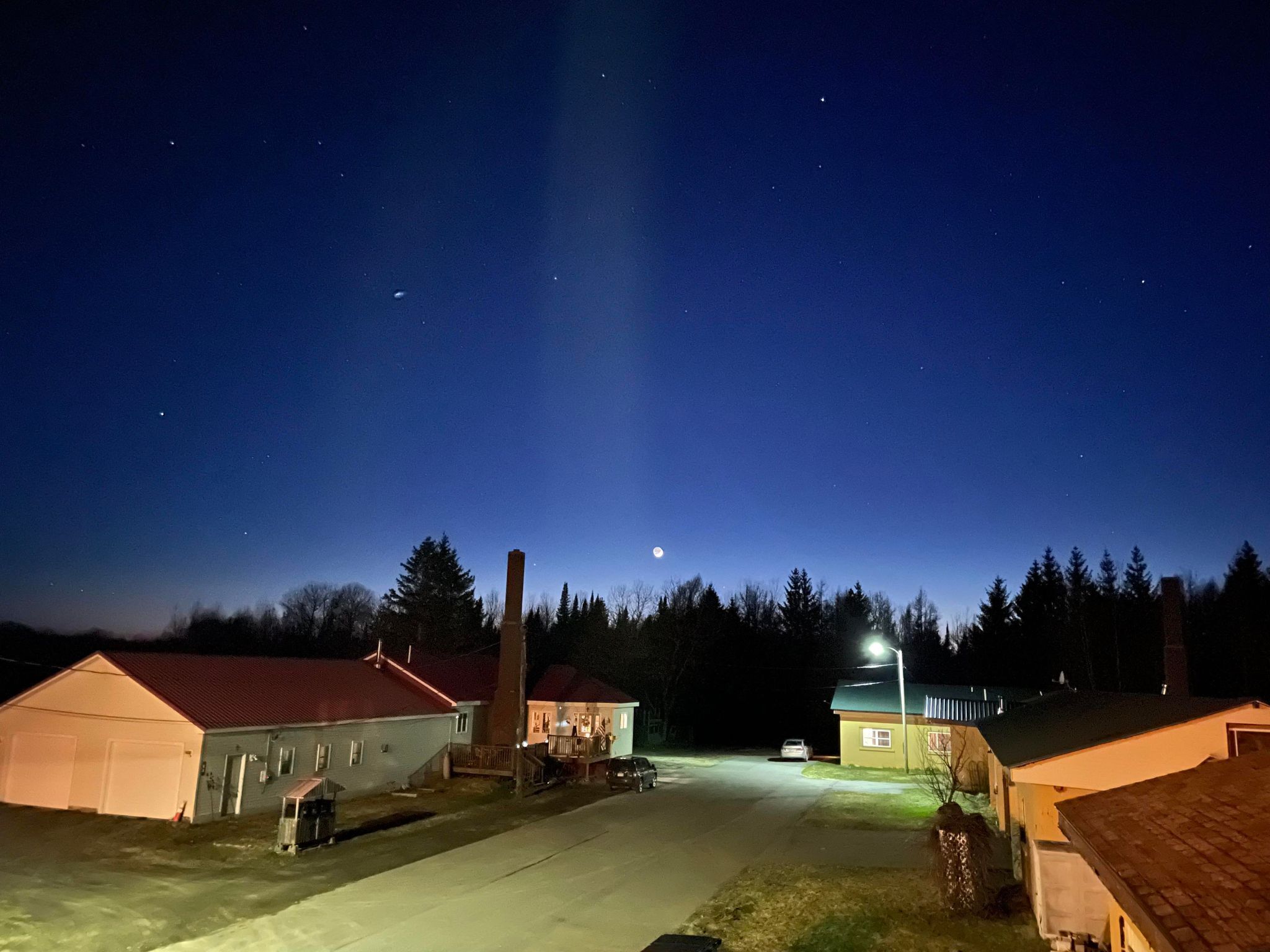 Ein ländliches Nachtbild mit klarem Himmel, einem sichtbaren Mond und einer Straße mit Gebäuden und Bäumen.