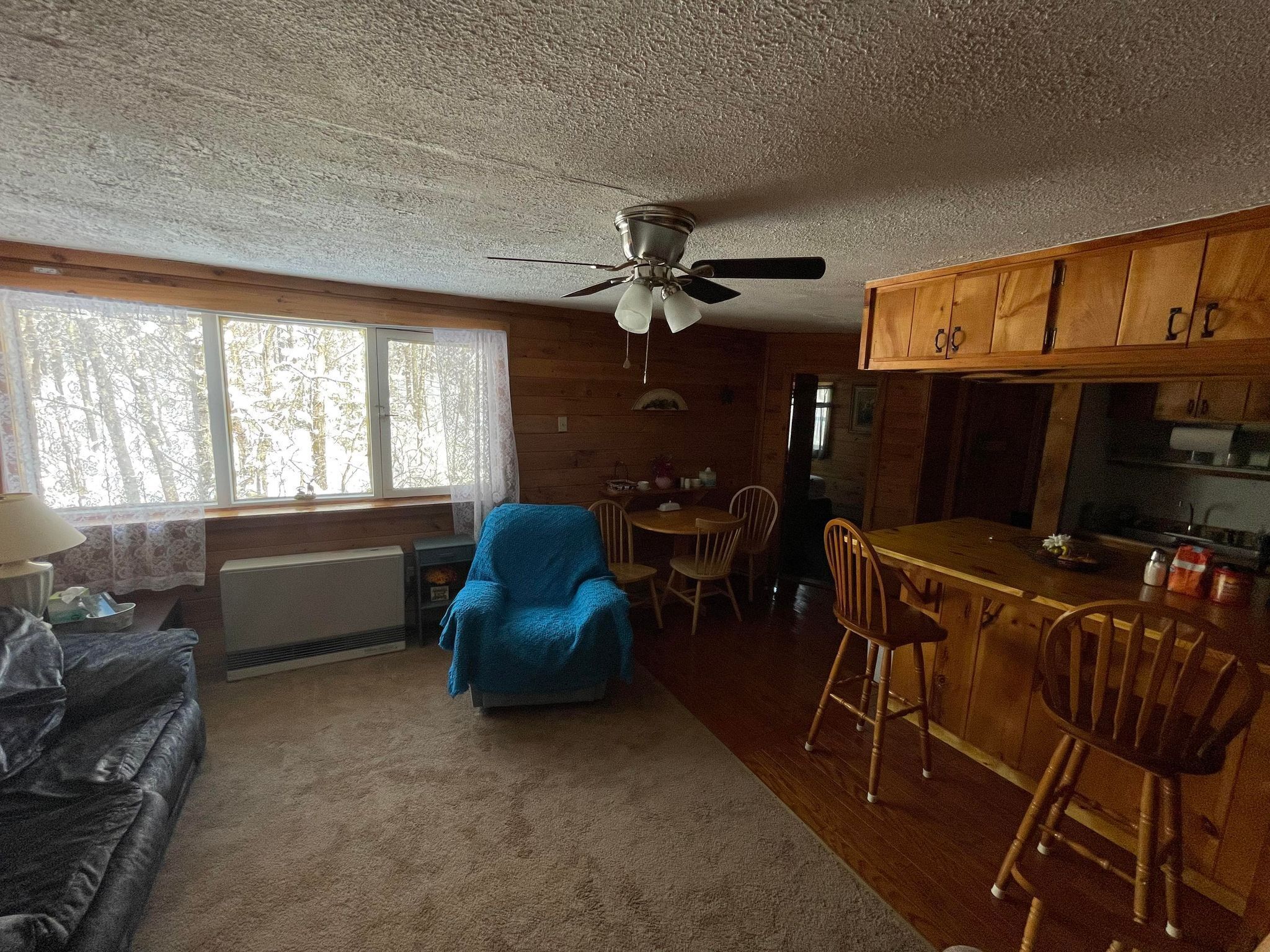 A cozy cabin interior with a dining area, a blue armchair, wooden cabinets, and a ceiling fan.