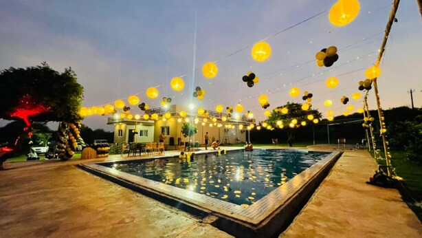 A swimming pool area with a house in the background, surrounded by string lights. - Gurugram Aravali Farmhouse  (Sohna)