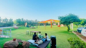 A grassy outdoor area with a picnic table, a large umbrella, and a few people relaxing. - Gurugram Aravali Farmhouse  (Sohna)