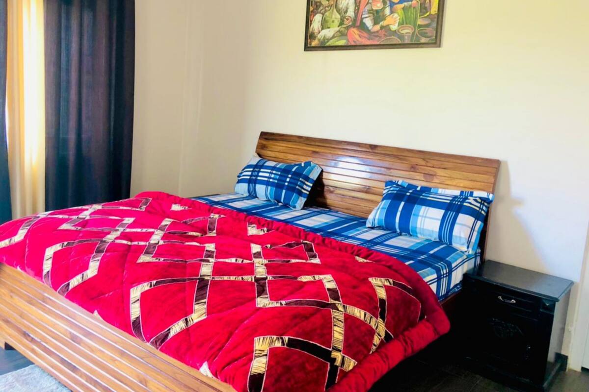 Un lit avec une couvre-lit à carreaux rouges et bleus, une tête de lit en bois et une table de chevet noire dotée d’un tiroir.
