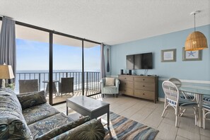 A spacious living area with a sofa, coffee table, and dining table. A TV is mounted on the wall, and there are framed pictures. - Carolina Reef 802 - Oceanfront, Pool, Hot Tub - Sleeps 4 (North Myrtle Beach)