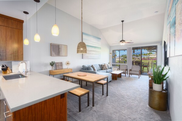 Eine moderne Küche mit Kochinsel, ein Essbereich mit Holztisch sowie ein Wohnzimmer mit Sofa und Sesseln.