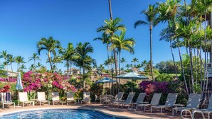 Ein Swimmingpool mit Liegestühlen und Sonnenschirmen, umgeben von Palmen und bunten Blumen.