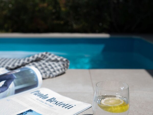 A glass of water with a lemon slice, a newspaper, and a towel by a pool.
