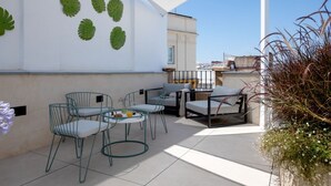 Une terrasse extérieure avec une table et des chaises, une petite table ronde avec une boisson, et des feuilles vertes décoratives sur le mur.