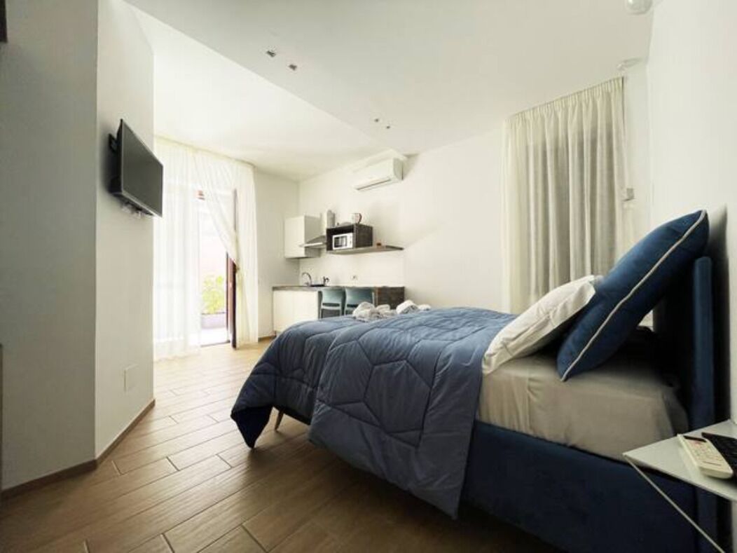 A modern hotel room with a bed, a desk, a TV, and a kitchenette.