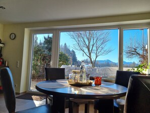 A dining area with a table and chairs, a clock on the wall, and a view of trees and mountains through large windows.