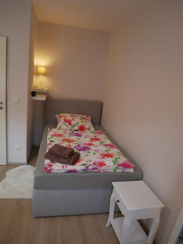 A small, neatly arranged bedroom with a single bed, a bedside table, a lamp, and a white rug.