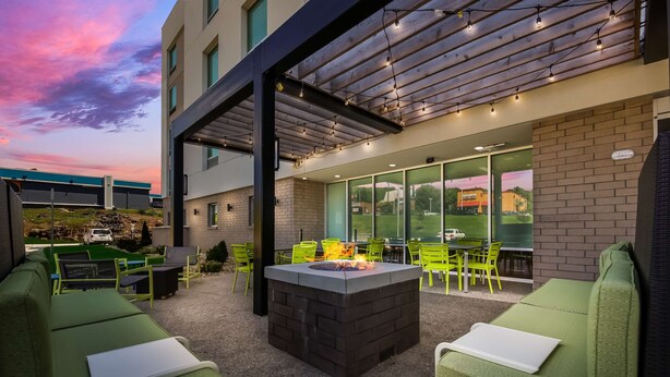 An outdoor seating area with green cushioned benches, a central fire feature, and a covered patio with string lights. - Home2 Suites By Hilton Cookeville (Cookeville)