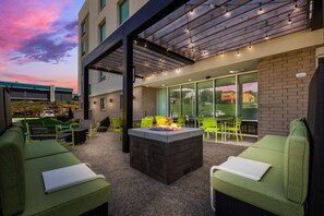 Un espacio al aire libre con bancos acolchados verdes, una chimenea central y un porche cubierto con luces colgantes.
