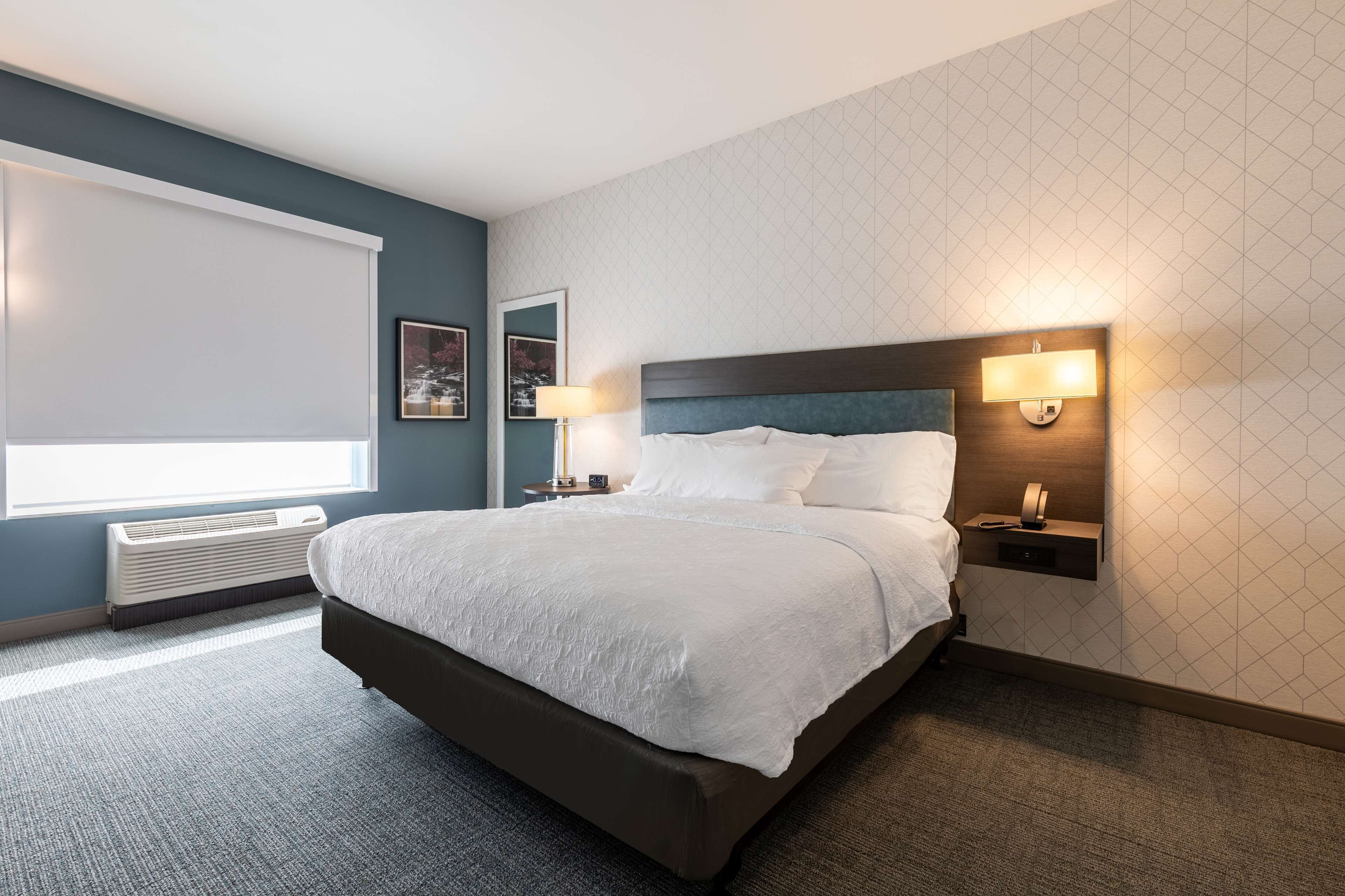 A hotel room with a large bed, a window with blinds, a wall-mounted heater, and a nightstand with a lamp.