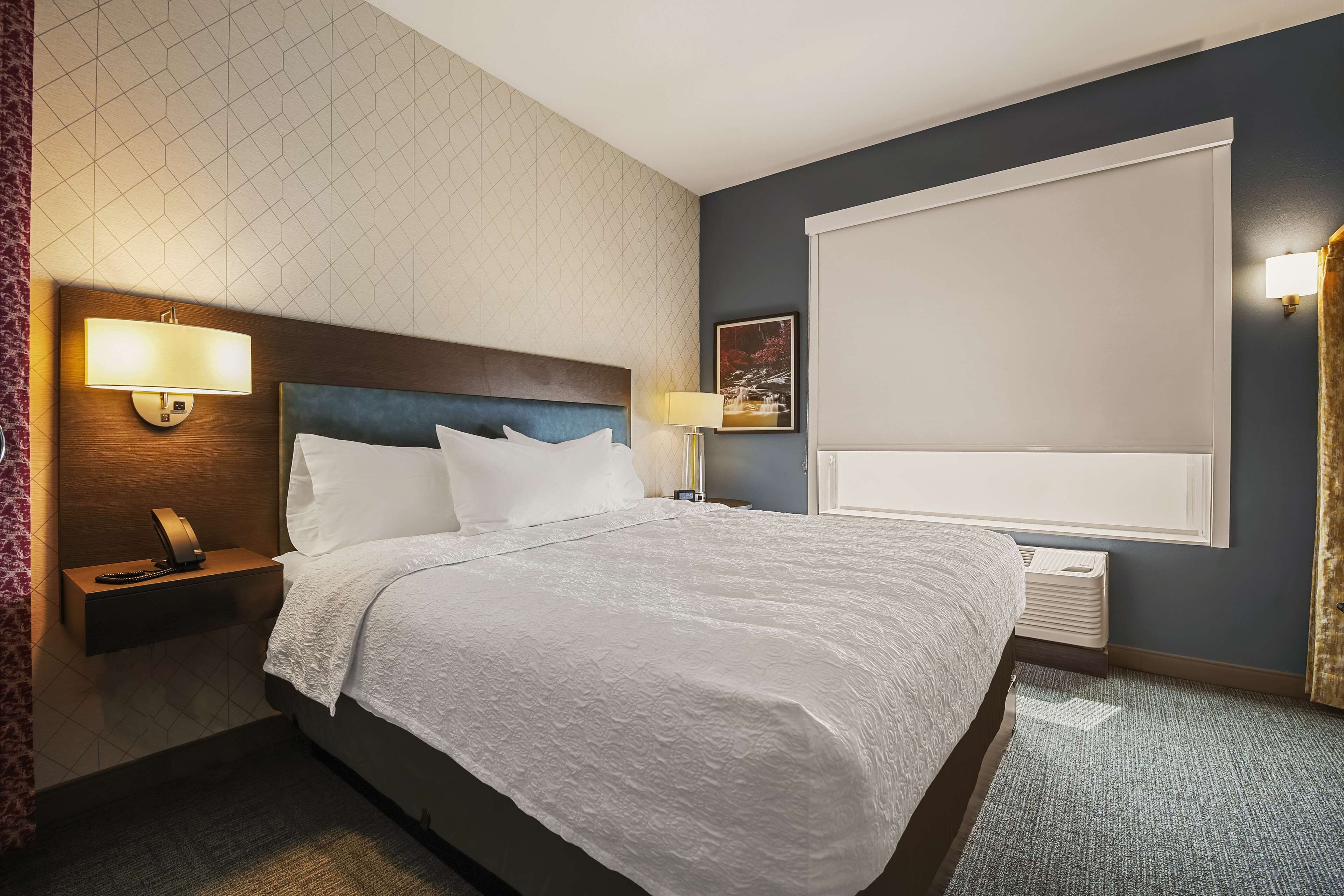 A hotel room with a large bed, a nightstand with a lamp, a wall-mounted TV, and a window with blinds.