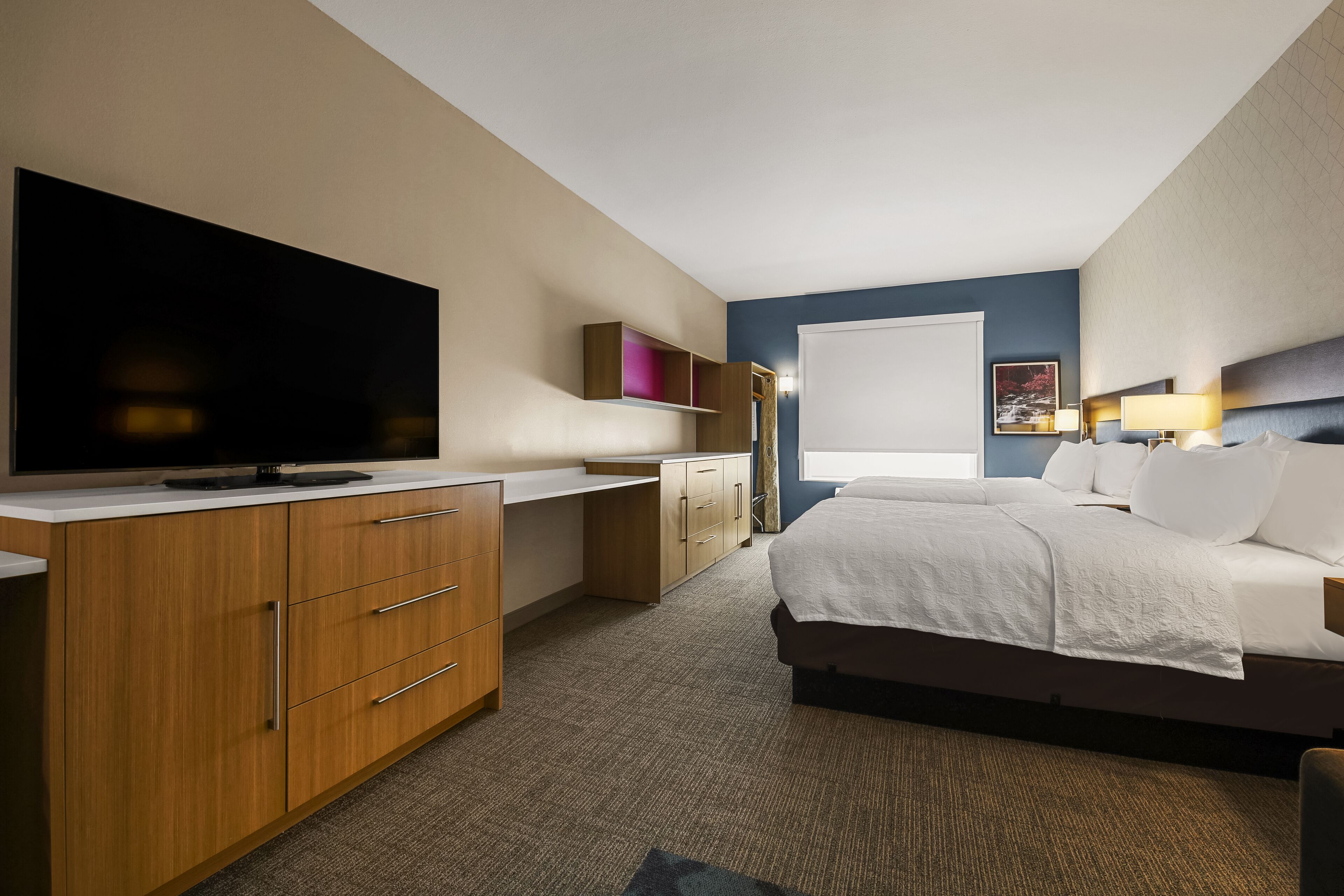 A hotel room with a large flat-screen TV, a bed with white linens, a desk with a computer, and a window with a view of the outdoors.