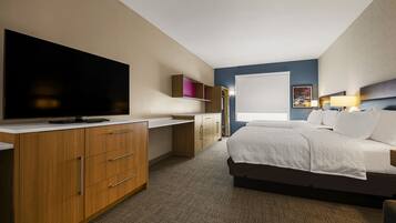 A hotel room with a large flat-screen TV, a bed with white linens, a desk with a computer, and a window with a view of the outdoors.