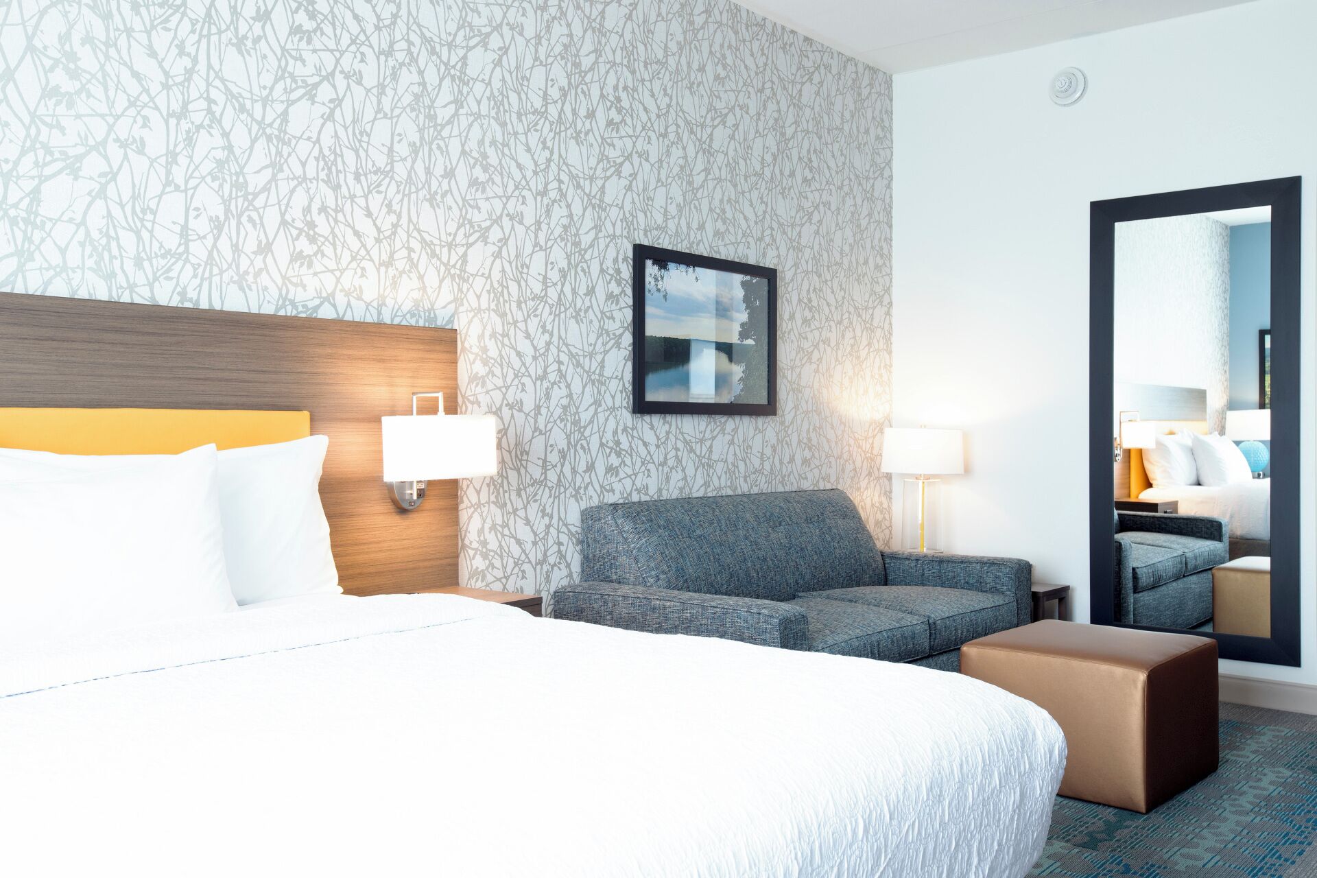 Habitación de hotel con una cama grande, un sofá gris, un pequeño otoman y un espejo que refleja otra habitación.