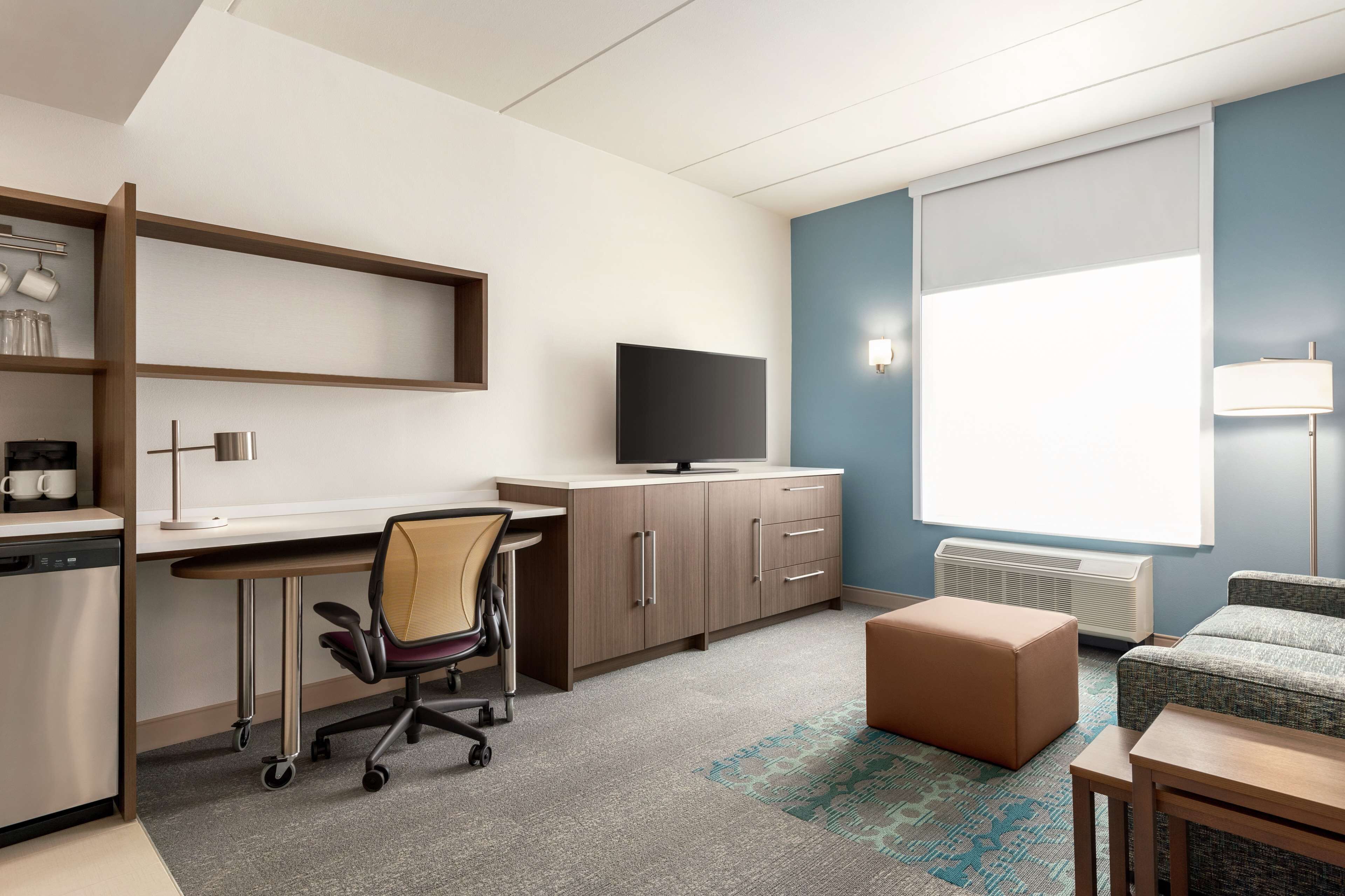 A modern hotel room with a kitchenette, a flat-screen TV, a sofa, and a large window with blinds.