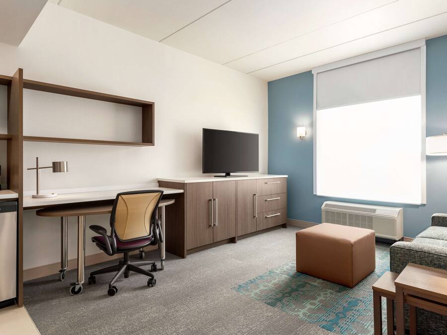 A modern hotel room with a kitchenette, a flat-screen TV, a sofa, and a large window with blinds.
