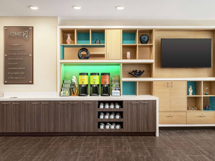 Uma área de recepção de hotel com design moderno, incluindo um balcão com máquinas de café, uma TV de tela plana e diversos objetos decorativos nas prateleiras.