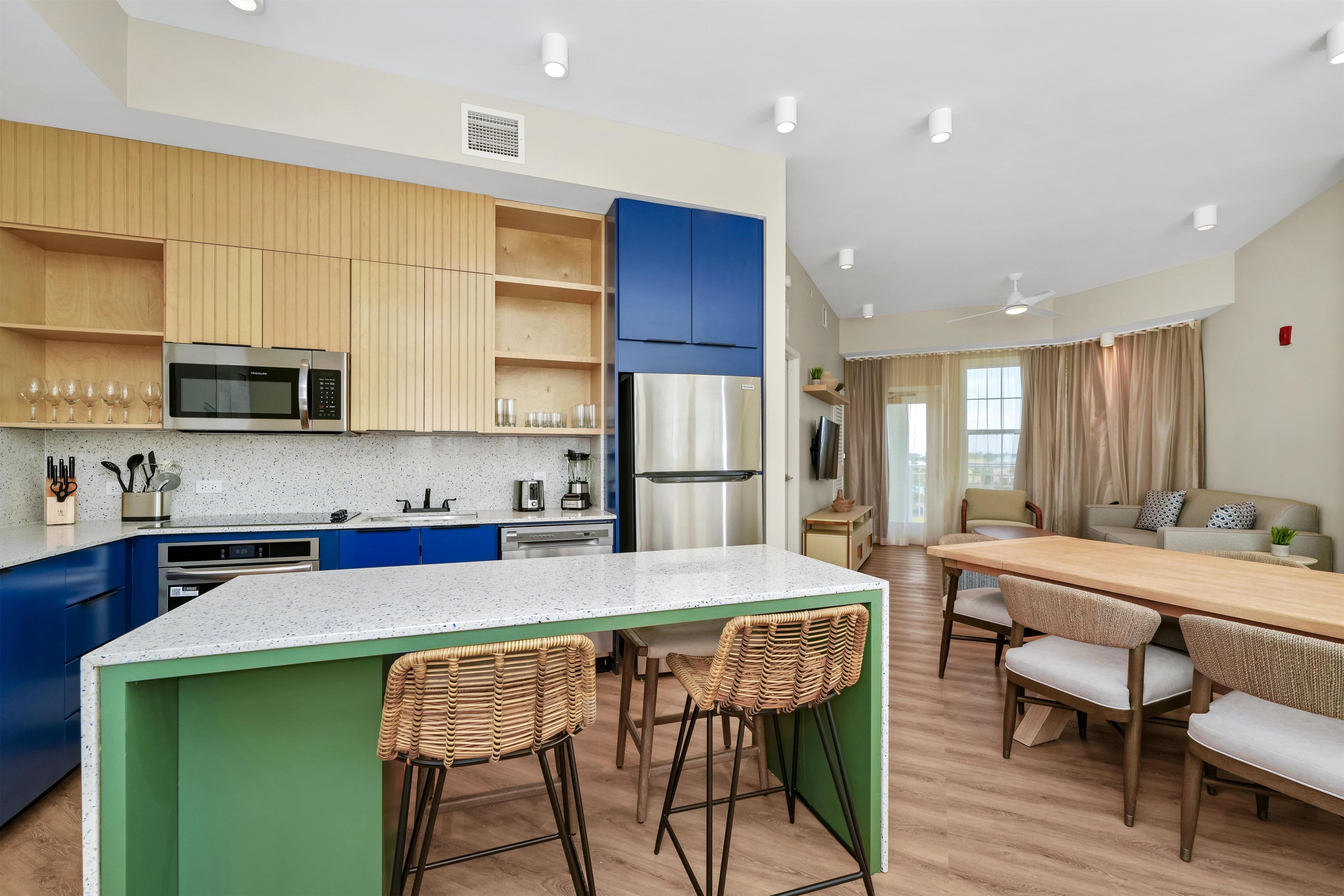 A modern kitchen with a central island, bar stools, a dining table, and a living area with a sofa and TV.