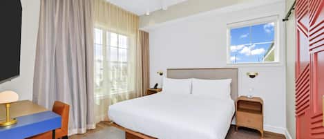 Una habitación de hotel con cama, escritorio, silla, mesita de noche, televisor y ventana con cortinas.