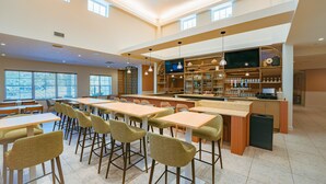 A spacious dining area with high tables and chairs, a bar area with shelves and a television, and large windows allowing natural light. - Embassy Suites By Hilton Orlando Sunset Walk (Kissimmee)