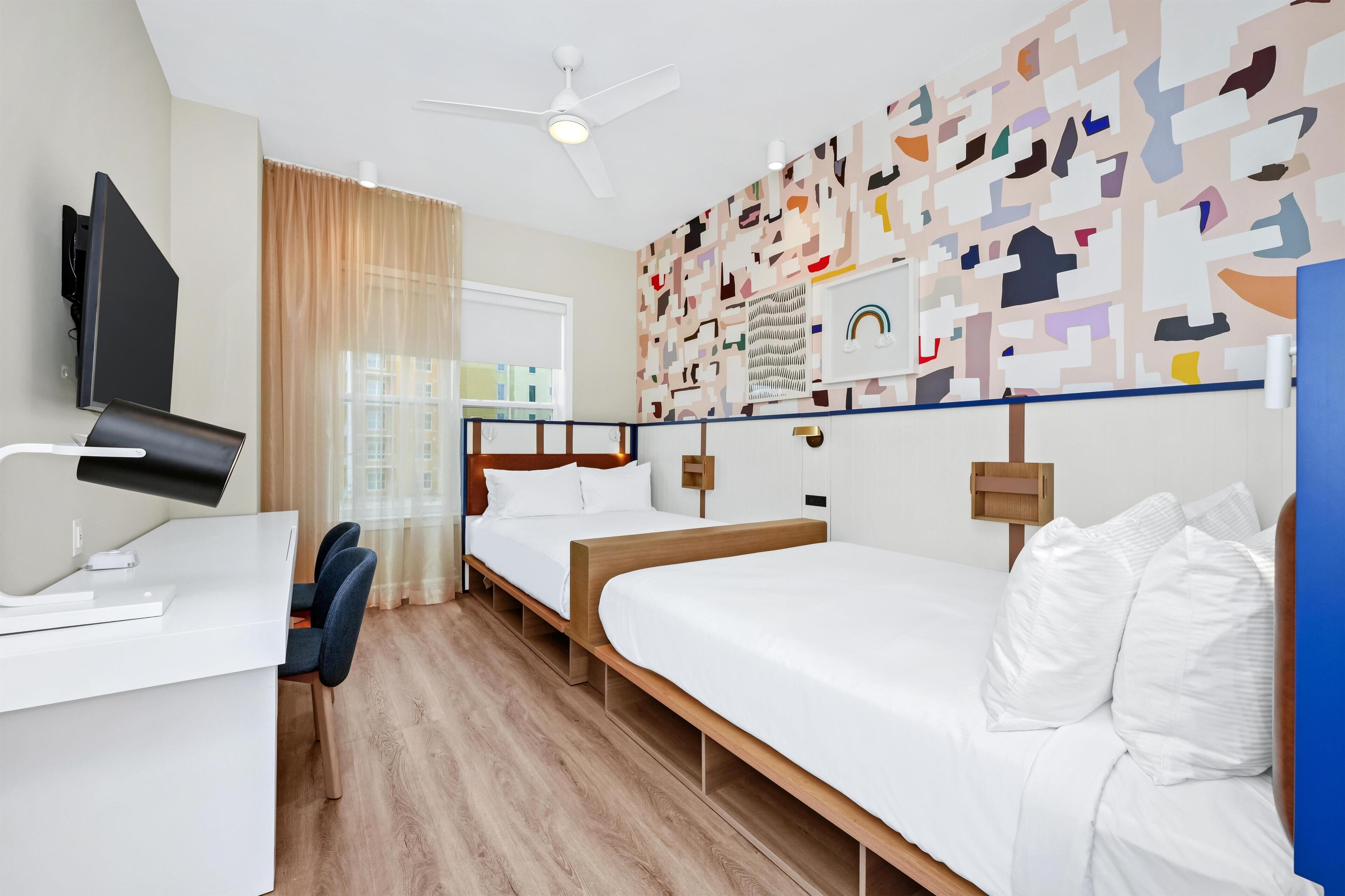 Hotellihuone, jossa on työpöytä, kaksi sänkyä, kattotuuletin, ikkuna verhoilla ja seinä, jossa on geometrinen kuvio.