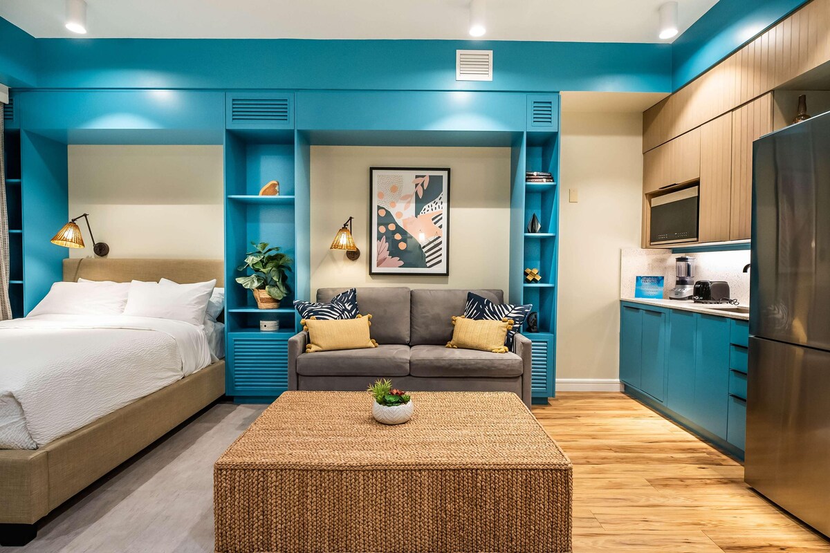 A modern room with a bed, sofa, kitchenette, and a painting on the wall.