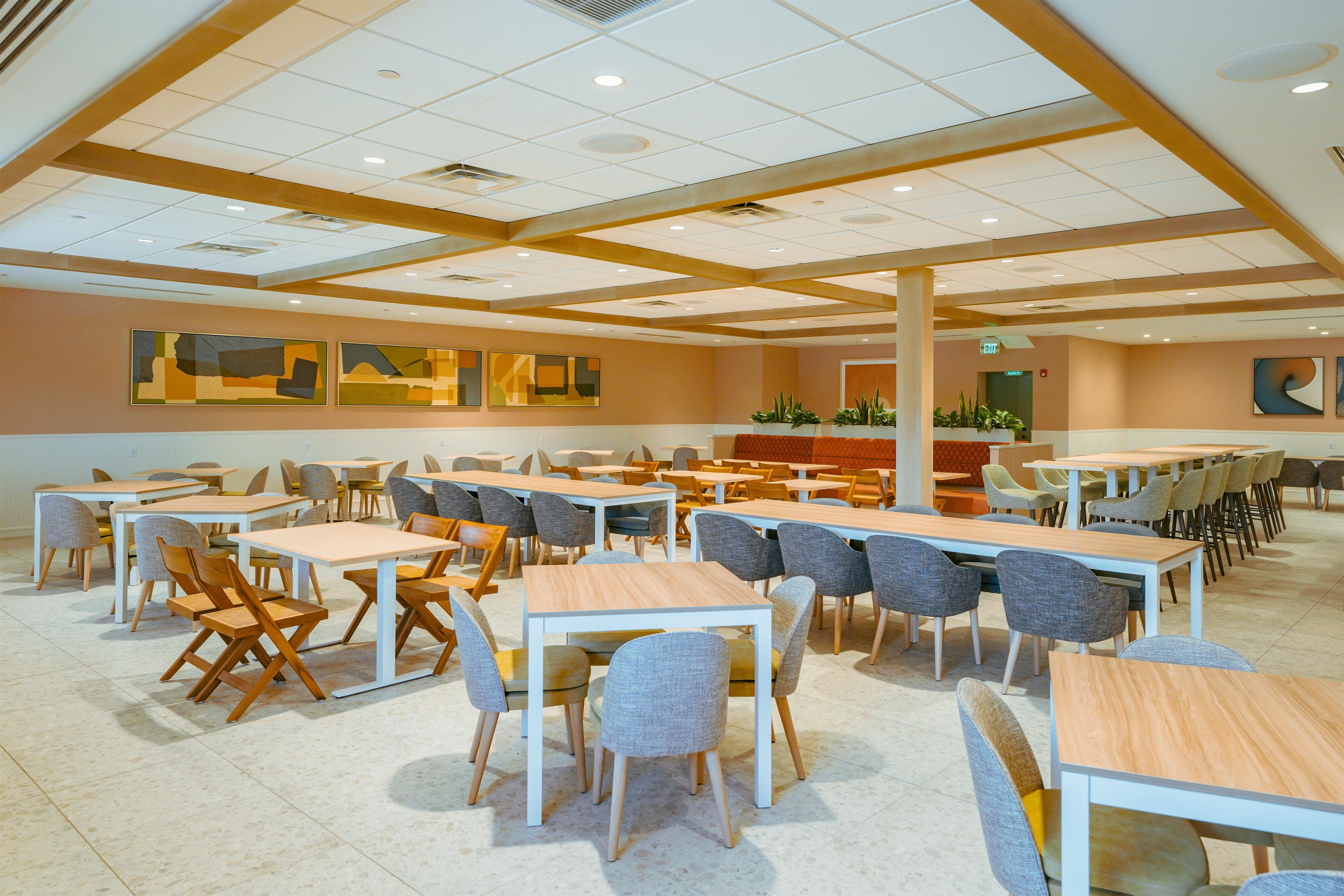 Un vaste espace dédié à la salle à manger, avec de nombreuses tables et chaises, un plafond haut doté d’un éclairage encastré et des œuvres d’art abstraites sur les murs.