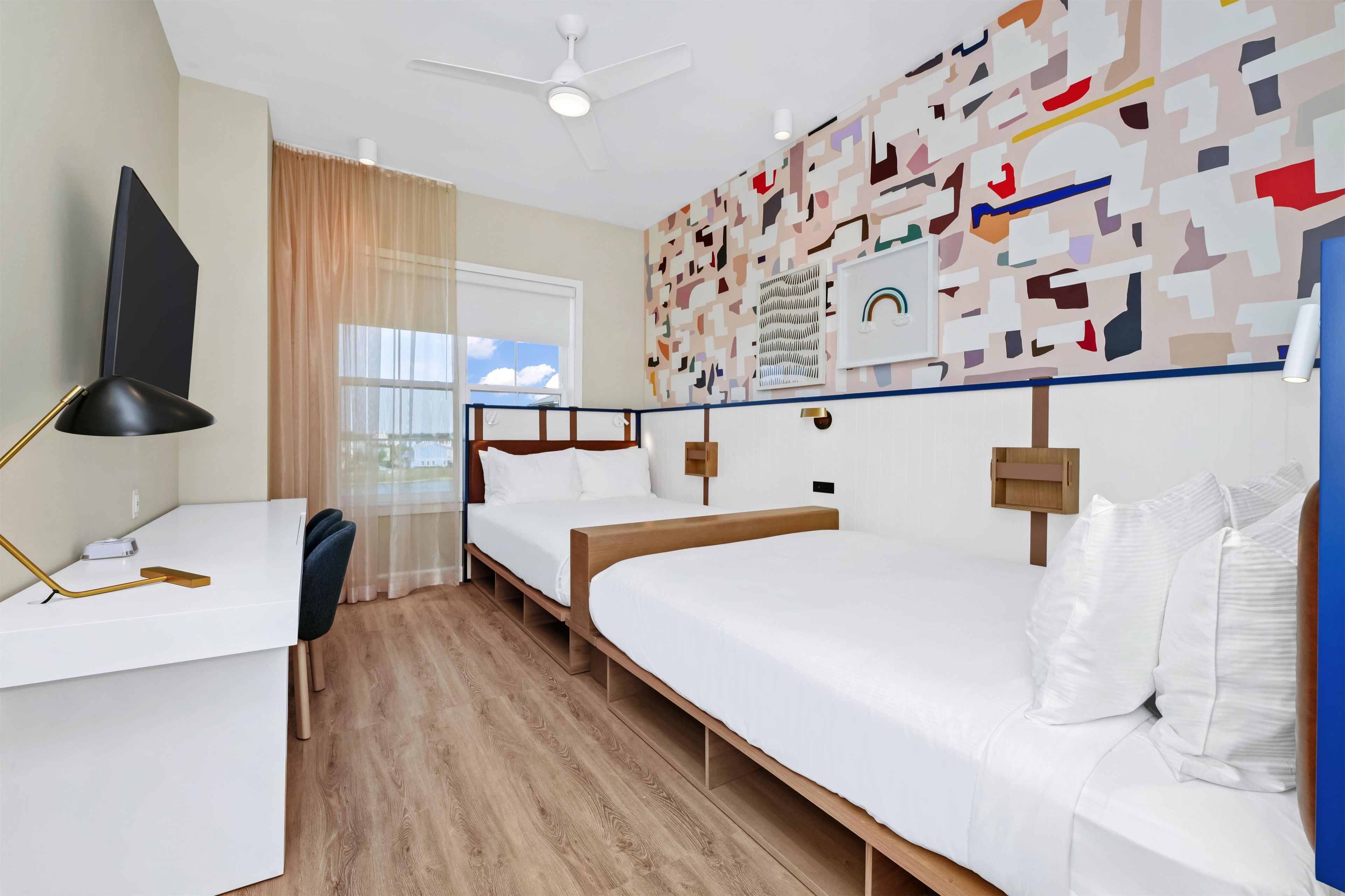 Habitación de hotel con escritorio, dos camas, ventilador de techo, ventana con cortinas y una pared con arte abstracto.
