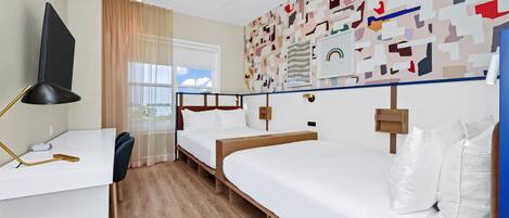 Habitación de hotel con escritorio, dos camas, ventilador de techo, ventana con cortinas y una pared con arte abstracto.