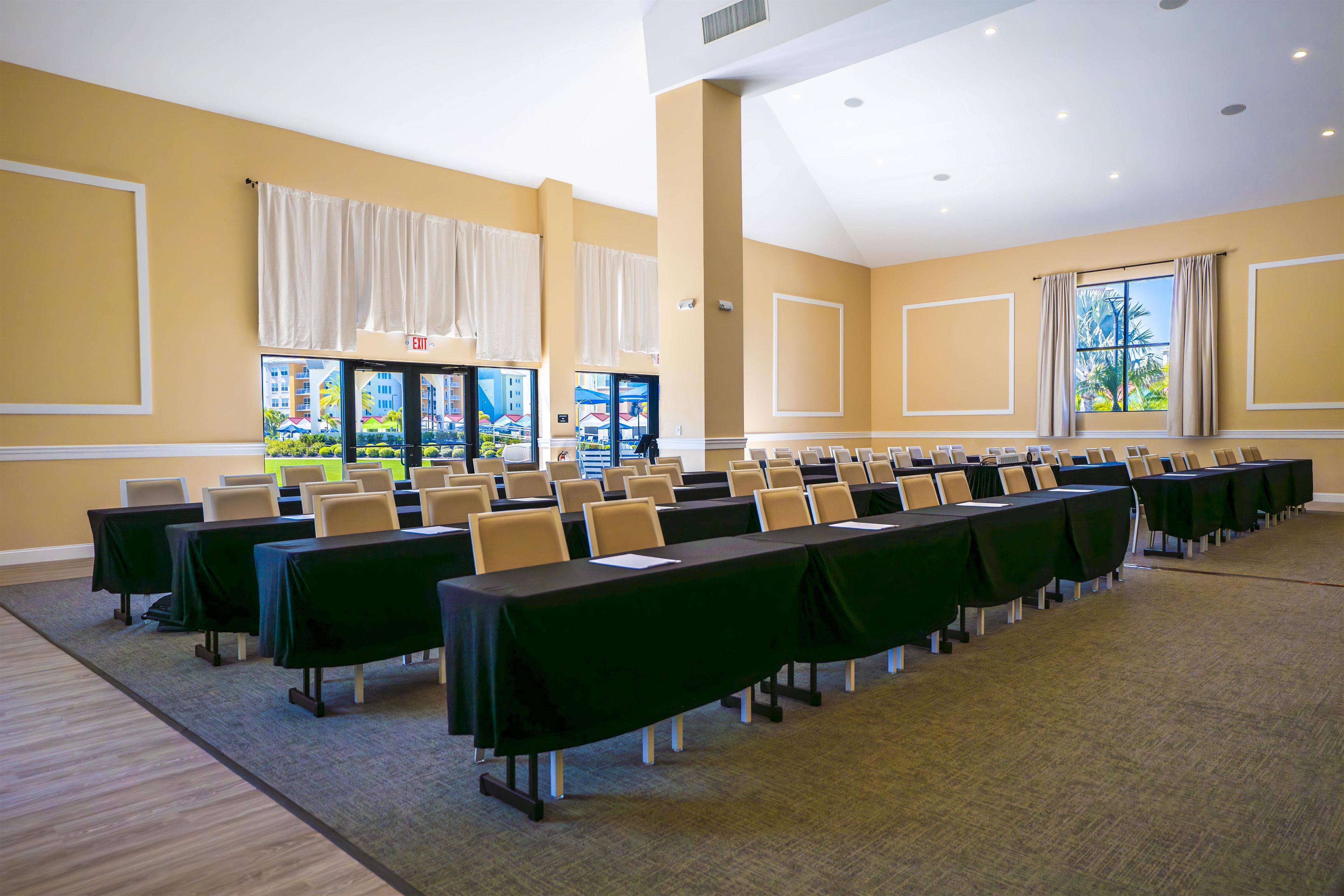 A conference room with rows of tables and chairs, large windows with curtains, and an exit sign.