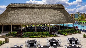 An outdoor dining area with round tables and chairs, a thatched roof structure, and a pool in the background. - Embassy Suites By Hilton Orlando Sunset Walk (Kissimmee)