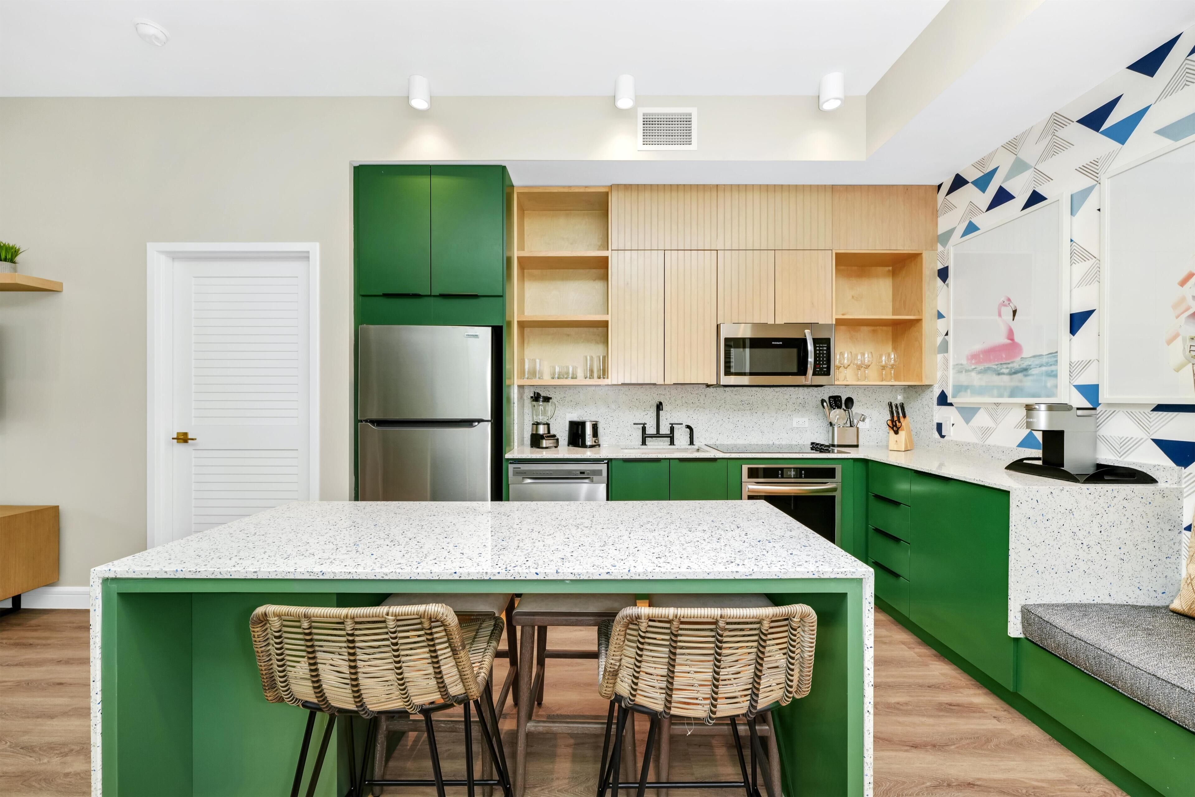 Una cocina moderna con armarios verdes, encimera blanca y dos taburetes de mimbre.
