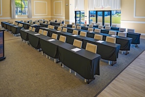Un salón de conferencias con filas de mesas y sillas, ventanas amplias y una puerta de salida.