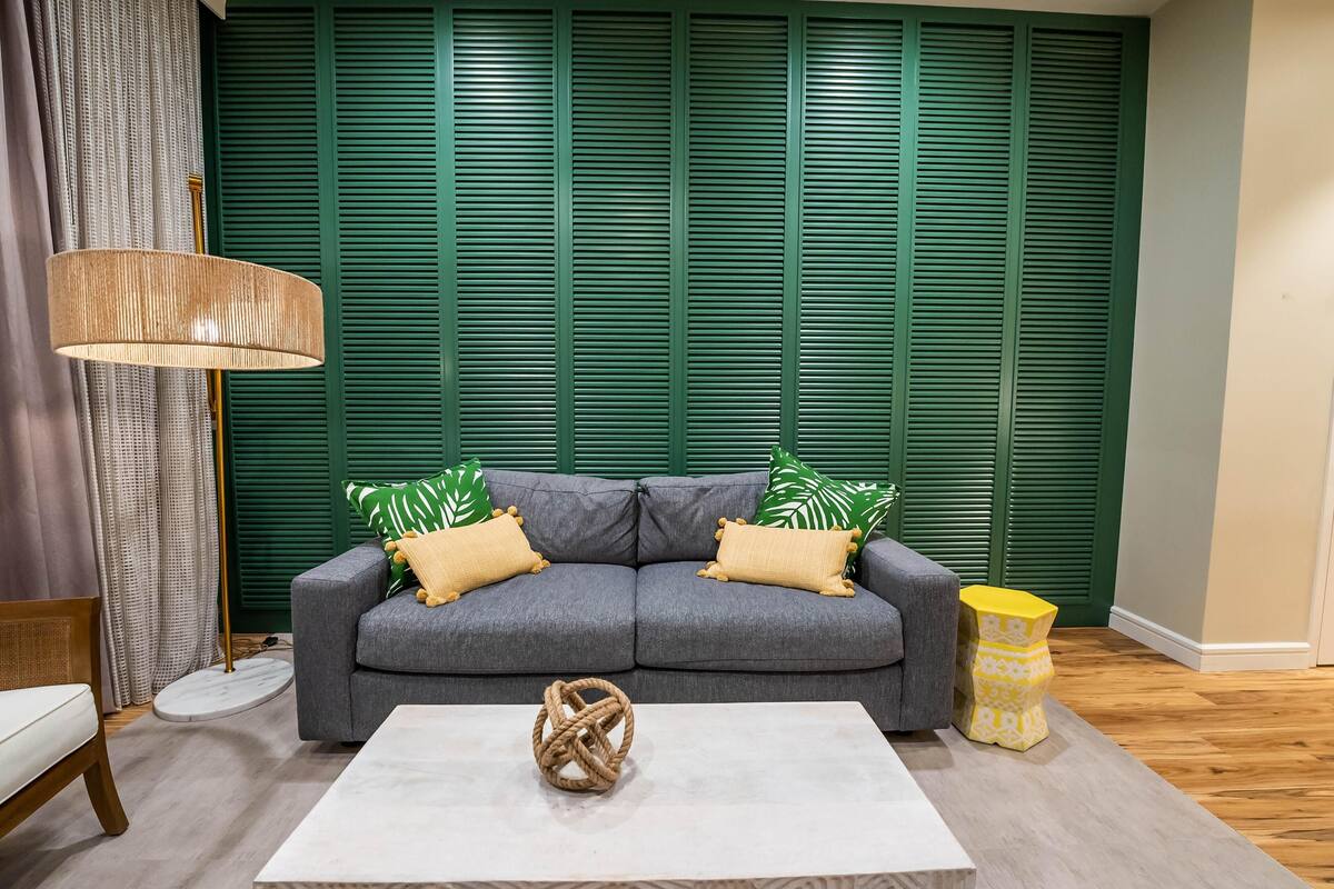 Een kamer met een grijze bank, een witte salontafel, een lamp en een groene muur met luiken.