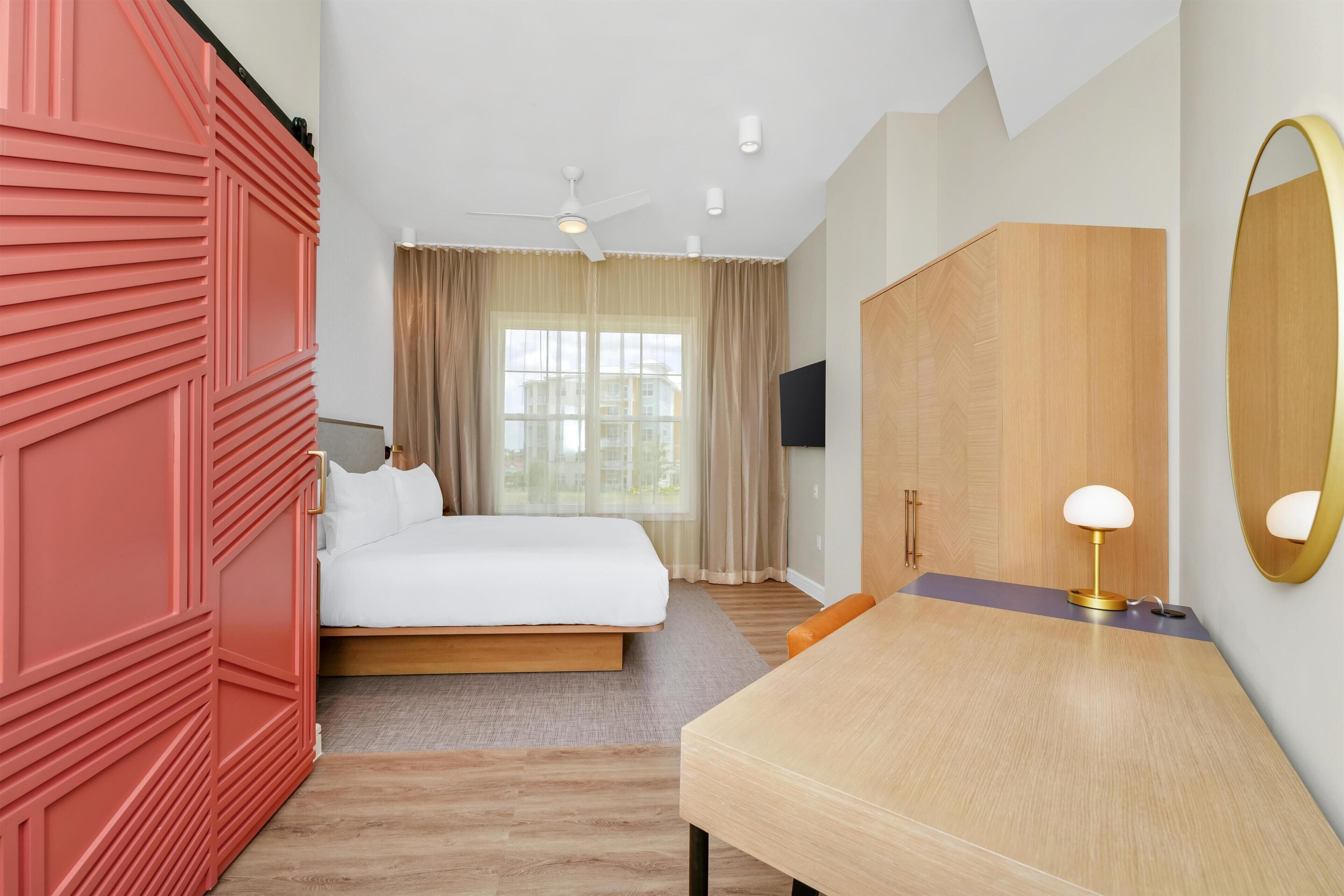A hotel room with a bed, a desk, a chair, a TV, a ceiling fan, and a large mirror.