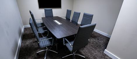 A conference room with a dark wooden table, six black chairs, a flat-screen TV mounted on the wall, and carpeted flooring.