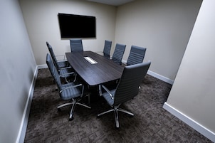 A conference room with a dark wooden table, six black chairs, a flat-screen TV mounted on the wall, and carpeted flooring.