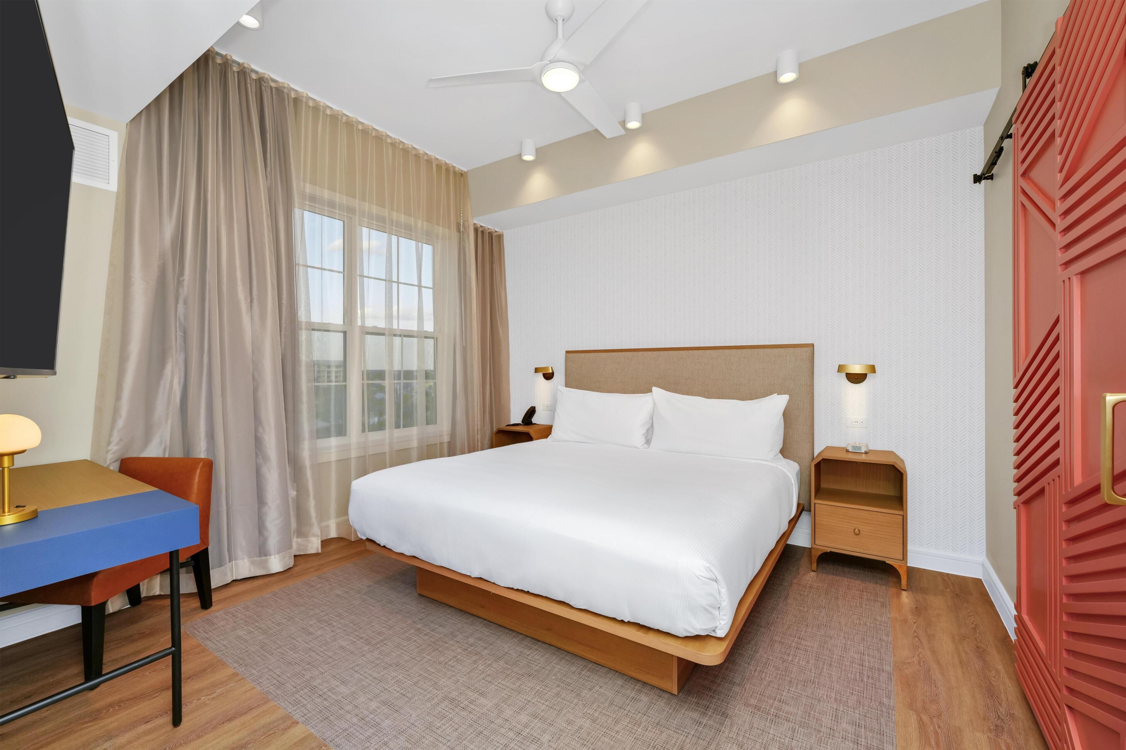 Habitación de hotel con una cama grande, un escritorio, una silla, una mesita de noche, un armario y un ventilador de techo.