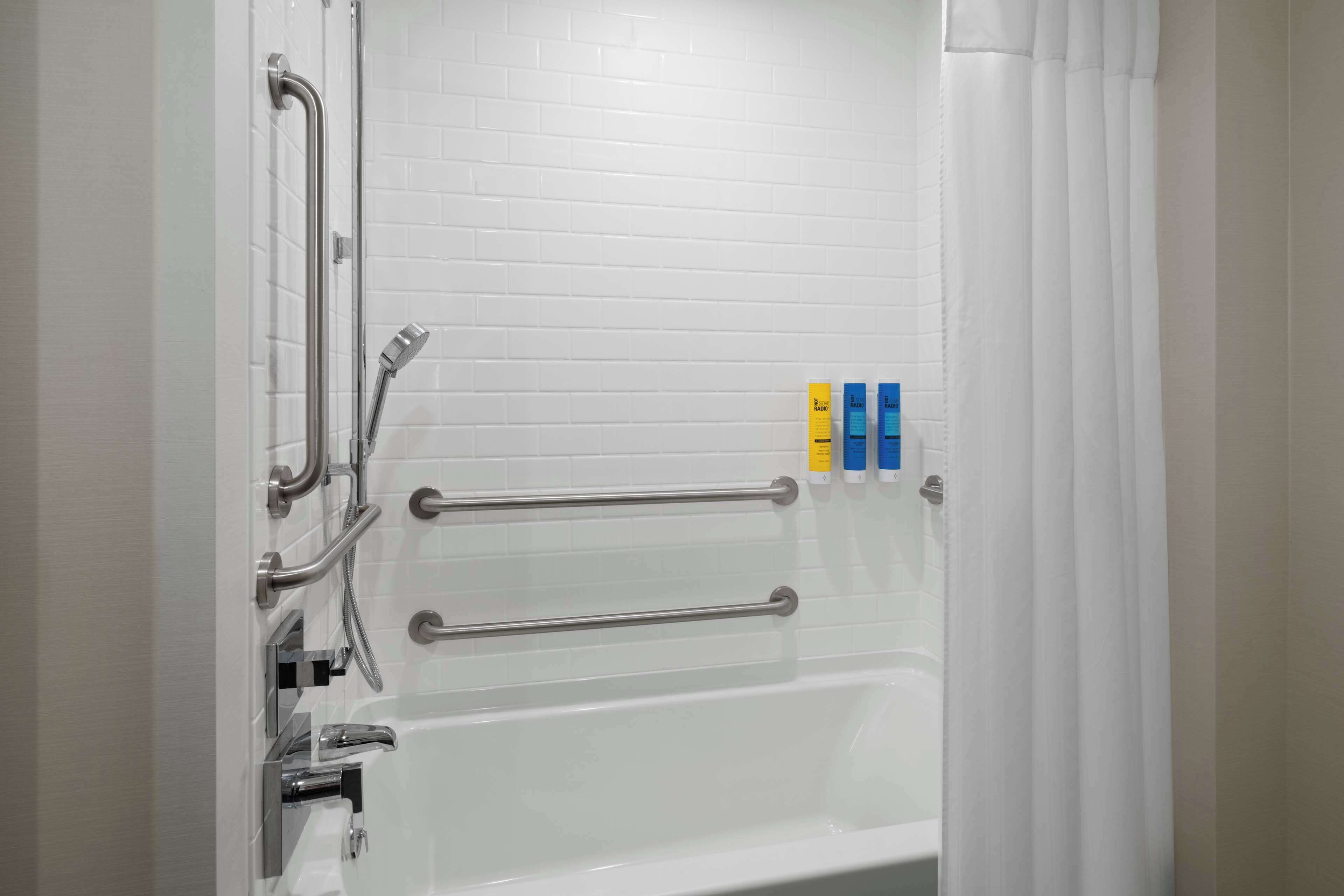 A bathroom with white tiled walls, a bathtub, and a shower with grab bars. There are three bottles of soap on the wall.