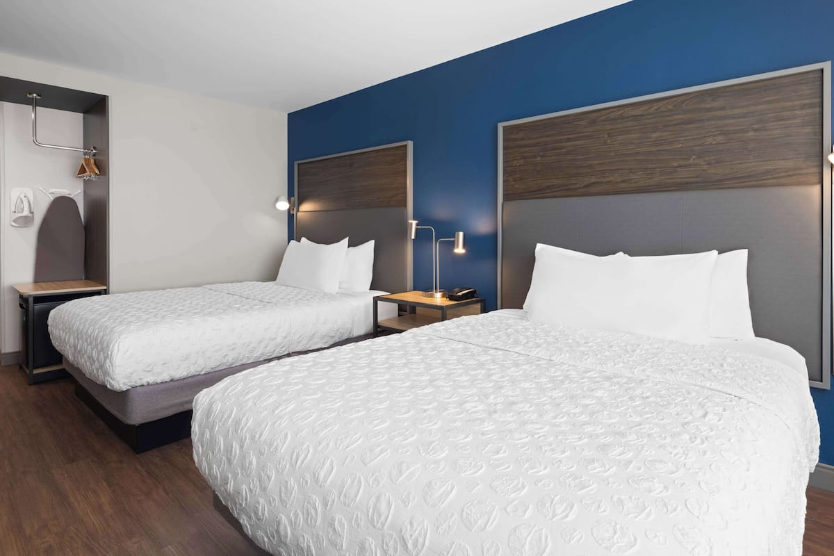 A hotel room with two single beds, a wooden headboard, a nightstand with a lamp, and a small table.