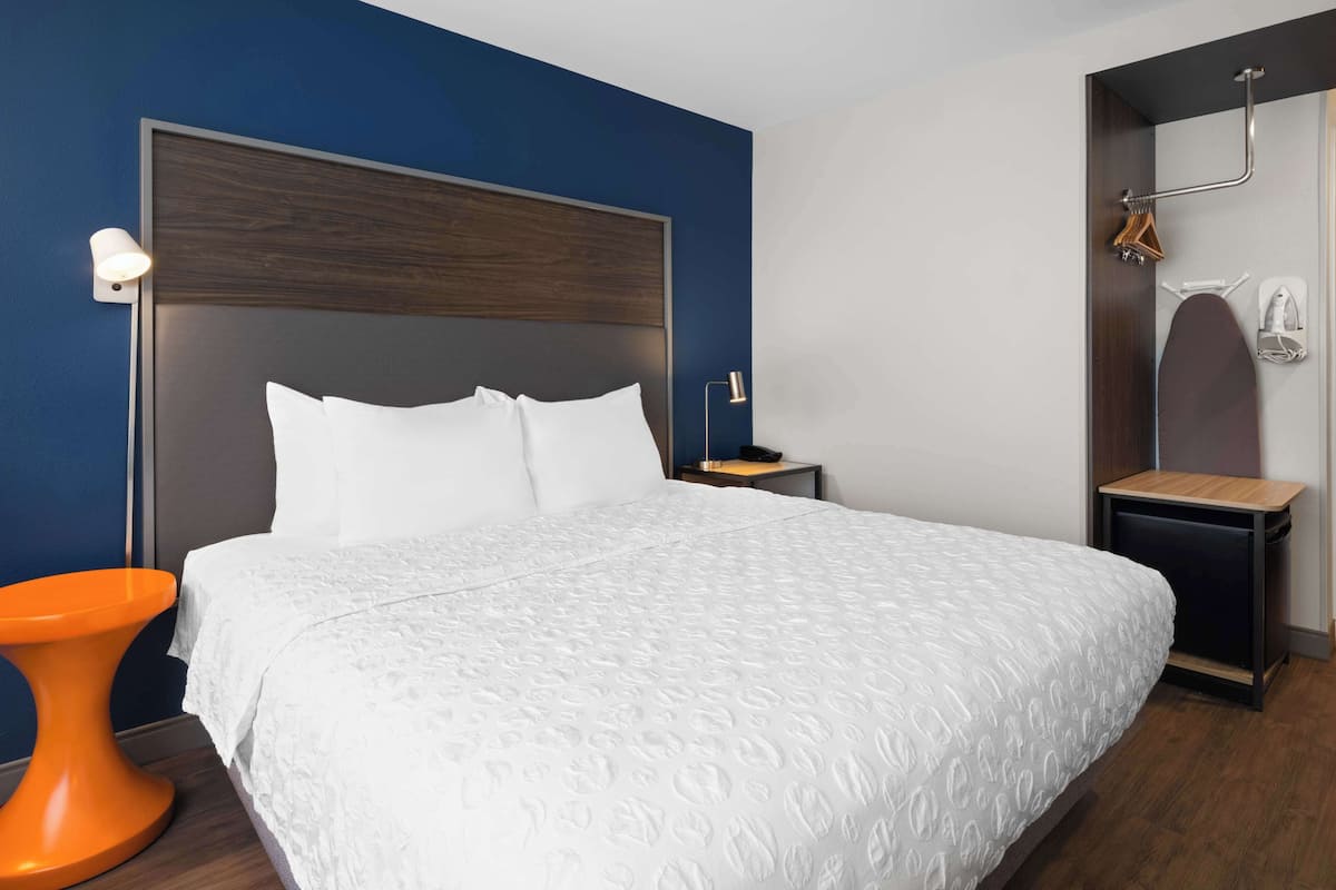 A neatly made bed with a white patterned bedspread, a wooden headboard, and a small orange stool beside it.