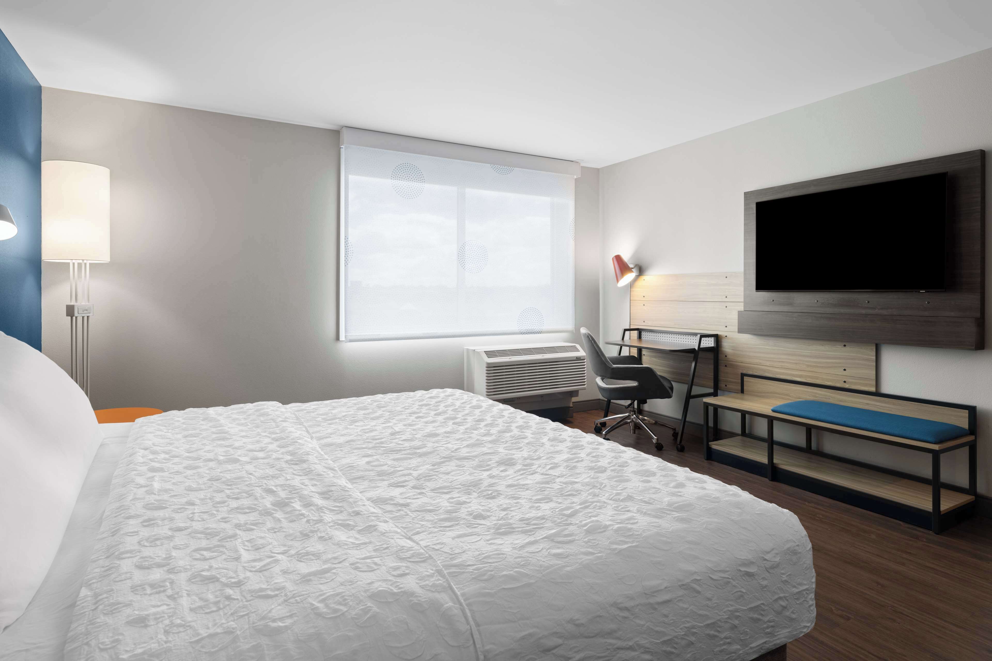 A hotel room with a large bed, a desk with a chair, a TV mounted on the wall, and a window with blinds.