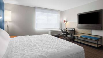A hotel room with a large bed, a desk with a chair, a TV mounted on the wall, and a window with blinds.