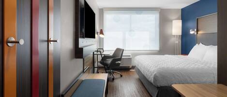 A hotel room with a bed, a desk with a chair, a bench, a TV, and a window with a view.