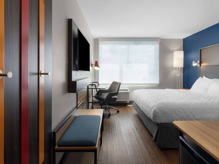 Habitación de hotel con cama, escritorio con silla, banca, televisor y ventana con vistas.